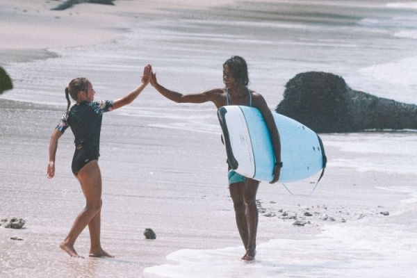 Two surfers high-five on a tropical beach, surrounded by palm trees and lush greenery, with one carrying a blue surfboard