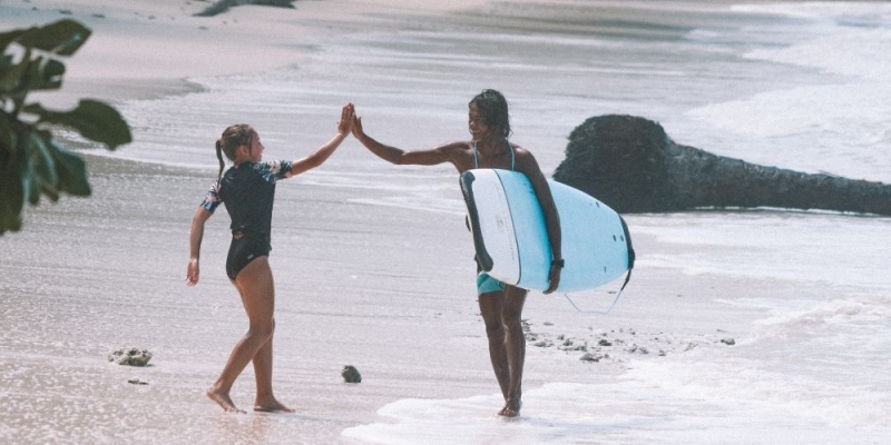 Two surfers high-five on a tropical beach, surrounded by palm trees and lush greenery, with one carrying a blue surfboard