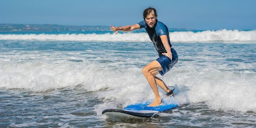 Young surfer standing on a surfboard, riding a small wave near the beach.